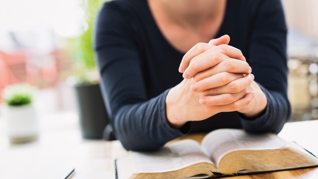 Bible study and faith concept - closeup shot of a woman holding her hands together and praying over an open Bible.